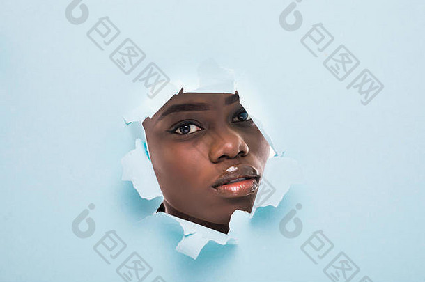 一个年轻美丽的非洲女孩的脸上有着明亮的妆容，嘴唇浮肿，凝视着蓝色纸上的一个洞。时尚、美容、化妆、化妆品、，