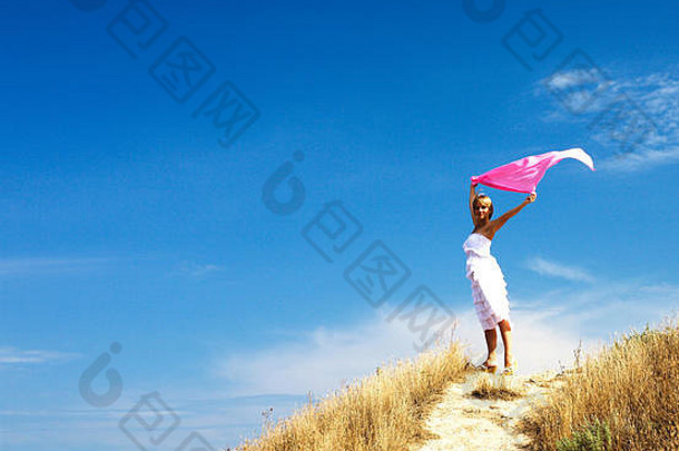 美丽的金发女人穿着白色衣服，戴着围巾站在海边的风中