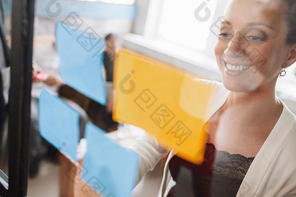 微笑年轻的业务女人坚持胶粘剂笔记玻璃墙把的想法玻璃墙