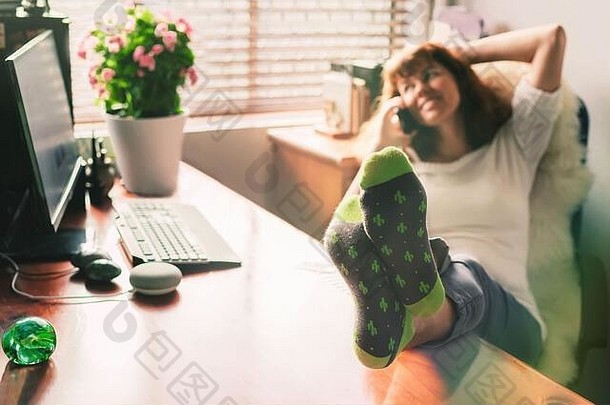 一位快乐的中年妇女坐在家里的办公室里，把腿放在桌面上，用手机说话。专注于脚。在家工作的概念。自我隔离。