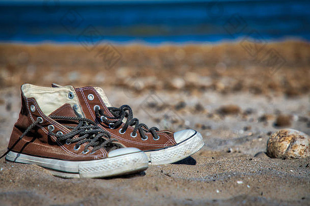 一双旧的棕色帆布风格复古<strong>篮球鞋</strong>（一只鞋聚焦，另一只鞋柔软聚焦）在沙滩上，水在沙滩上