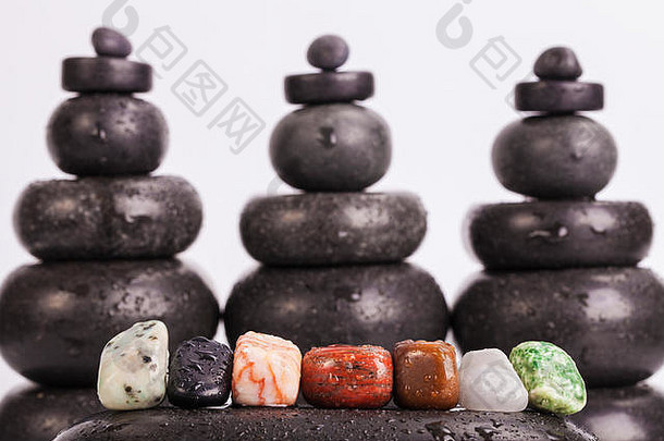 一排七个脉轮颜色的水晶排列在湿滑抛光的热按摩黑石上，背景模糊，由热按摩黑石制成