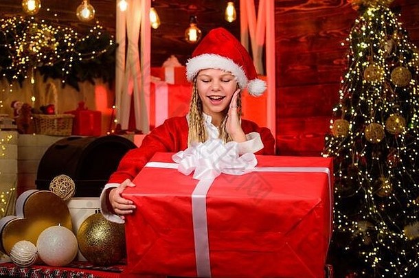 圣诞老人就在附近。用欢乐装饰家。圣诞节的心情。家庭节日庆典。新年快乐。戴着红色圣诞帽的孩子。拿着礼物盒的孩子。冬季购物大减价。圣诞树旁快乐的小女孩。