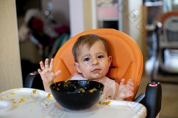 一个可爱的小女孩坐在一盘食物前的橙色儿童座<strong>椅</strong>上，她举起双手看着摄像机。到处都是食物残渣。特写镜头