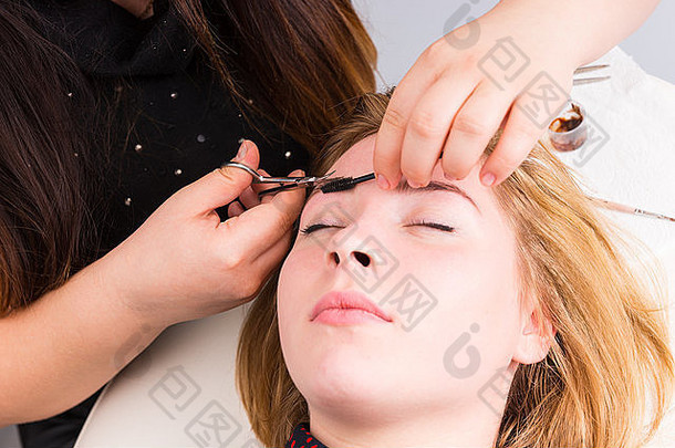 美容师在沙龙水疗中心用剪刀和睫毛刷修剪眉毛、闭着眼睛躺着的金发女郎的特写镜头。