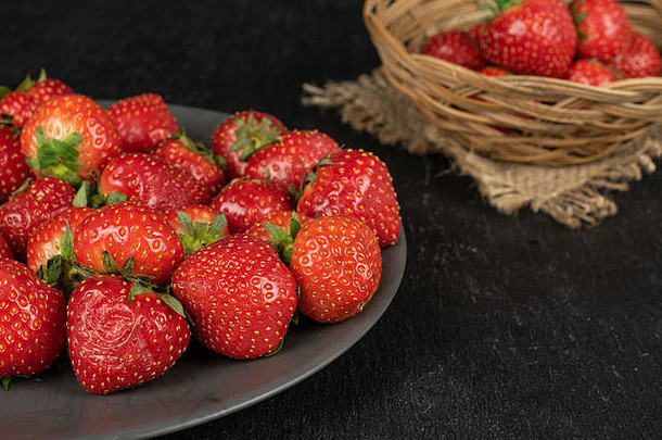 整批新鲜的红色草莓放在灰色陶瓷盘上，放在圆形藤碗中，黄麻织物放在灰色石头上