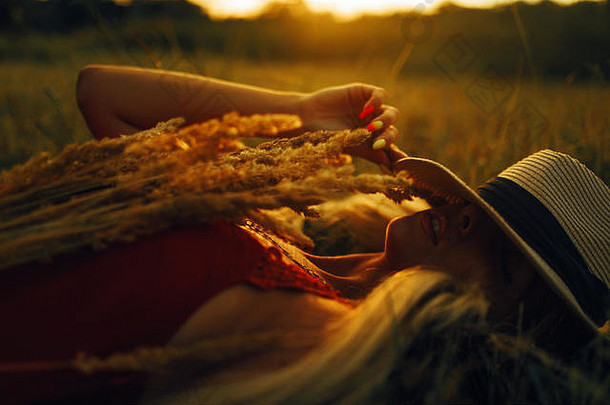 日落时分，戴着帽子的金发女郎躺在草地上的草丛中。