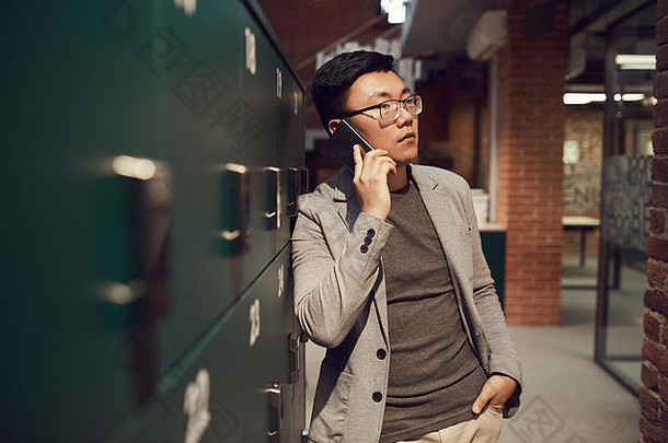 成功的亚洲商人站在现代办公室和复印空间的储物柜旁，通过智能手机讲话的腰身肖像