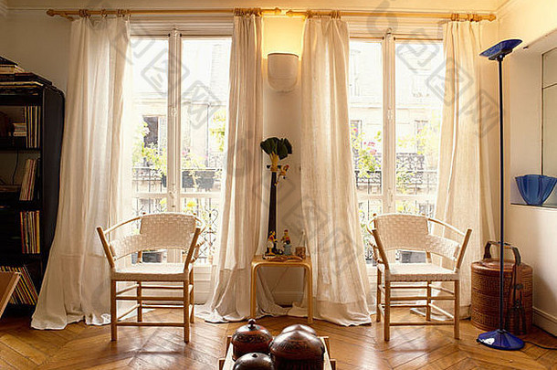 在铺有实木地板的起居室中，高双窗前有白色座椅和木制框架的椅子，白色窗帘