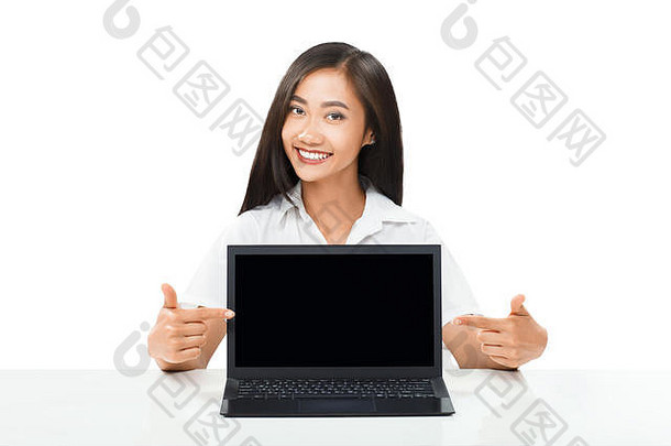 亚洲美女指着笔记本电脑显示器的肖像。年轻的女商人坐在桌旁，微笑着，展示着一台带有空白屏幕的便携式电脑。