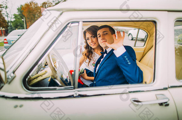 坐在老式汽车里的幸福新婚夫妇。新郎向服务员挥手致意