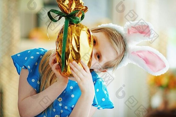 在阳光明媚的春日里，穿着波尔卡圆点蓝色整体和复活节兔子耳朵的现代儿童在现代客厅里，从包裹在<strong>金箔纸</strong>上的大耳朵向外张望