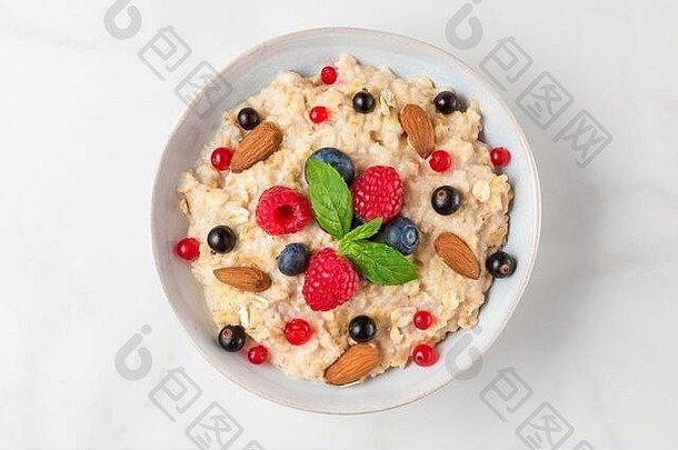 一碗燕麦粥，配新鲜浆果、杏仁和薄荷，在白色餐桌上享用健康饮食早餐。俯视图