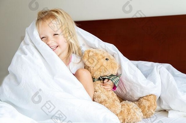 可爱的金发小女孩和玩具熊坐在床上。快乐的童年。COVID-2019冠状病毒疾病的家中滞留