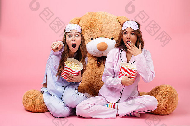 两个穿着睡衣的漂亮女孩正坐在一只大泰迪熊旁边，一边吃着粉色后背上的爆米花，一边用手指着镜头，吓得魂不附体