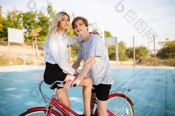 骑在自行车上的金发碧眼的漂亮男孩和女孩在篮球场上愉快地看着镜头。一对年轻可爱的情侣骑在自行车上