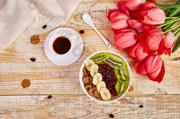 咖啡杯和碗，上面有格兰诺拉麦片、香蕉、猕猴桃、浆果和粉色郁金香花束，放在质朴的木桌上，母亲节或母亲节的早餐