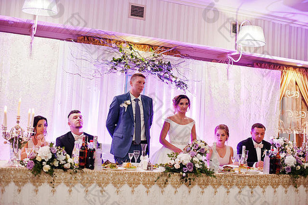 美丽的新婚夫妇站在桌旁，伴娘和伴郎坐在他们旁边。