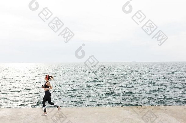 穿着深灰色运动上衣和带耳机的紧身裤的年轻女子在令人惊叹的海景背景下若有所思地奔跑
