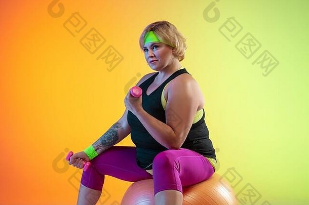 年轻的白人加大体型女模特在霓虹灯下的<strong>渐变橙色</strong>背景上训练。做举重锻炼。体育观念、健康生活方式、身体积极、平等。