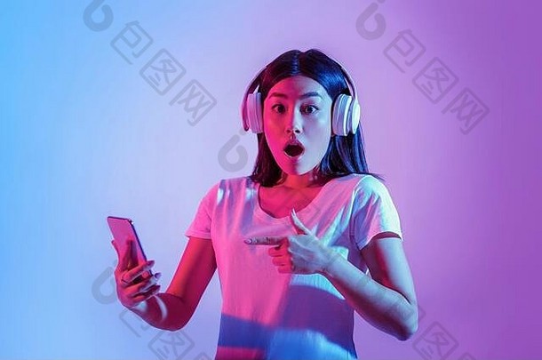 戴着大型无线耳机的亚洲女孩大吃一惊，用手指指着智能手机