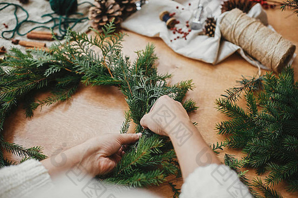 制作质朴的圣诞花环。手拿着杉木树枝、松果、线、浆果、剪刀放在木桌上。圣诞花圈工作坊。认证