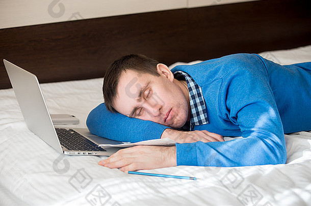 一幅随意迷人的年轻男子躺在床上，睡在笔记本电脑前，困倦、疲惫、过度工作或懒惰的肖像