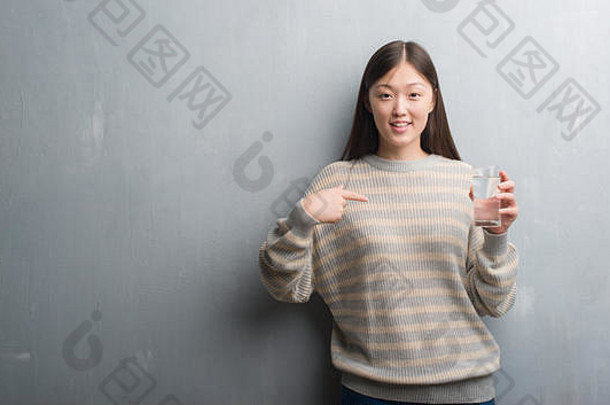 一位年轻<strong>的</strong>中国妇女在灰墙边喝水，惊讶地用手指着自己