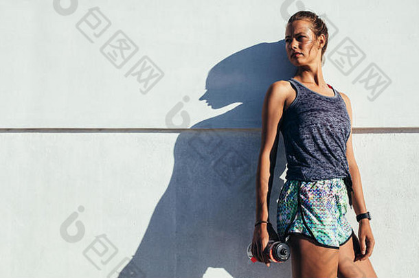 站在户外的年轻女子的肖像，水瓶对着<strong>墙</strong>向外看。女跑步者在训练后放松。