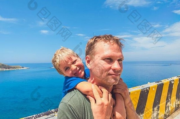 男孩坐在父亲的肩膀上笑。皮肤上没有photoshop防晒霜。海，云，岛屿背景。有趣的照片，快乐的生活方式，父亲的