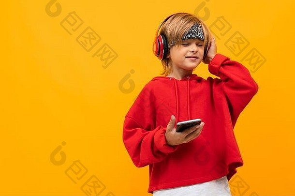 身着红色连帽衫、面带微笑的欧洲金发男孩戴着红色耳机专心听音乐，并将智能手机放在<strong>黄色</strong>墙上