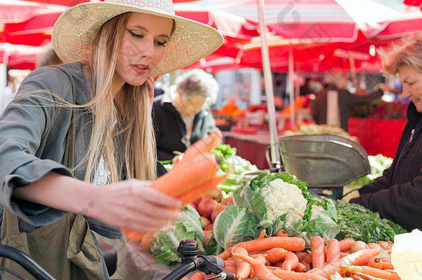 漂亮的金发女孩戴着草帽在市场上买蔬菜。