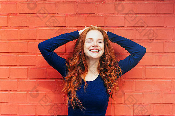 一位面带微笑的女士，身穿深蓝色长袖衬衫，双手枕在头后，靠在红砖墙上
