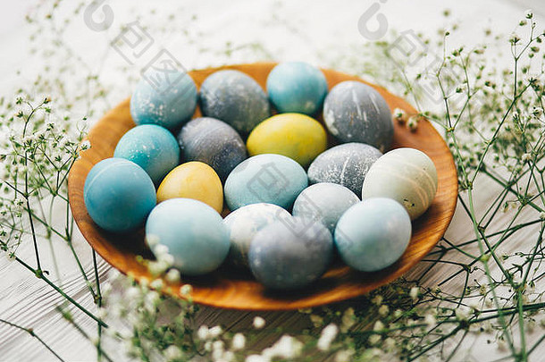 白色木质背景上的木制盘子上有春天花朵的时尚复活节彩蛋。现代复活节彩蛋用蓝色、灰色、黄色的天然染料绘制