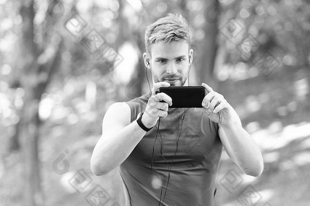 男子运动员集中面部拍摄智能手机照片自然背景。运动员训练计步器和耳机配件。运动员鬃毛健身跟踪器和智能手机。捕捉愉快的时刻。