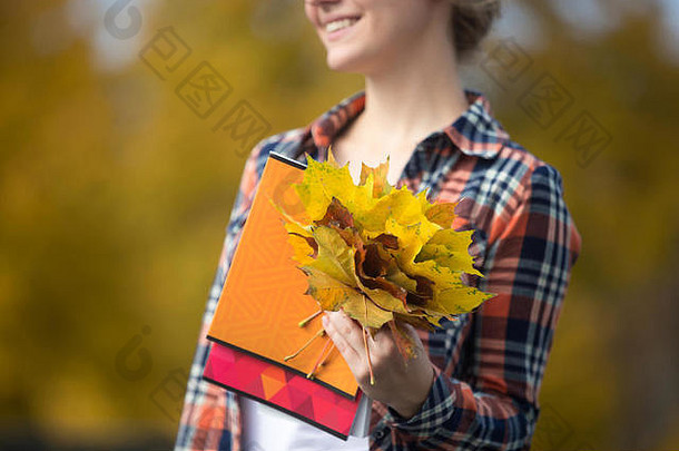 微笑女年轻的学生在户外持有黄色的叶子