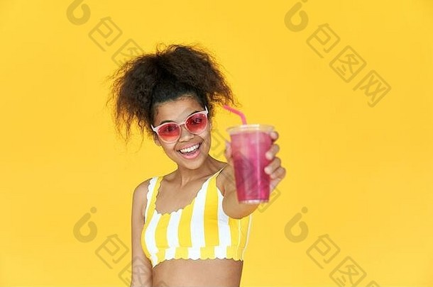 快乐的非洲女孩戴着墨镜拿着饮料看着黄色摄影棚里的摄像机。