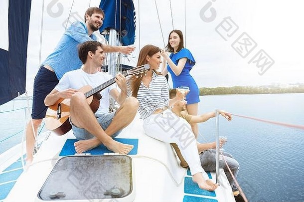 一群快乐的朋友在户外的船上聚会上喝着伏特加鸡尾酒，兴高采烈。年轻人在海上玩吉他旅游，有青春和暑假的概念。喝酒、度假、休息。