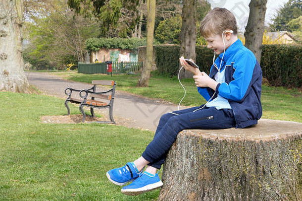 青少年男孩聪明的电话听会说话的坐着树桩英国公园少年社会媒体概念