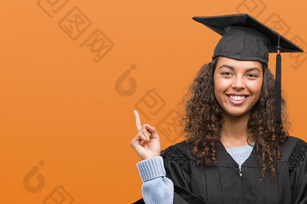 年轻的拉美裔女人穿毕业统一的快乐指出手手指一边
