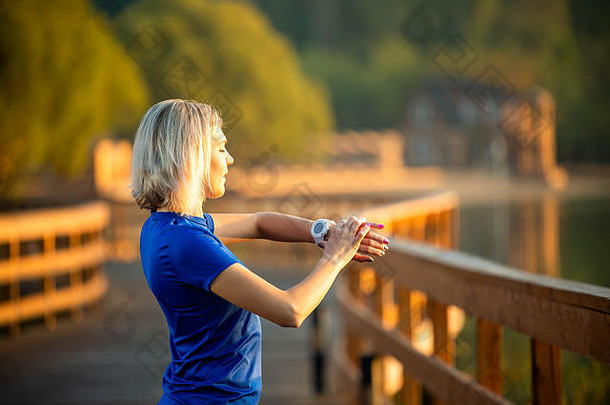 图为夏天，一位运动型女士站在公园的木桥上，看着手边的时钟。