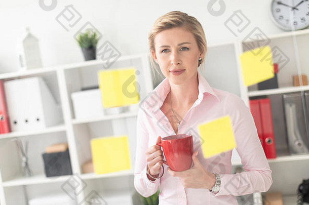 一个年轻的金发女孩站在办公室里，旁边是一块透明的<strong>贴纸板</strong>，手里拿着一个红色的杯子。