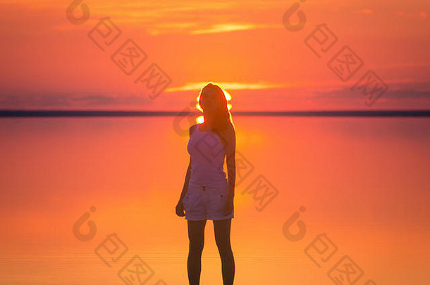 美丽的女模特站在海边地平线后的太阳前。盐湖埃尔顿平静的海水在日落时折射出女人的轮廓。那个女孩独自一人