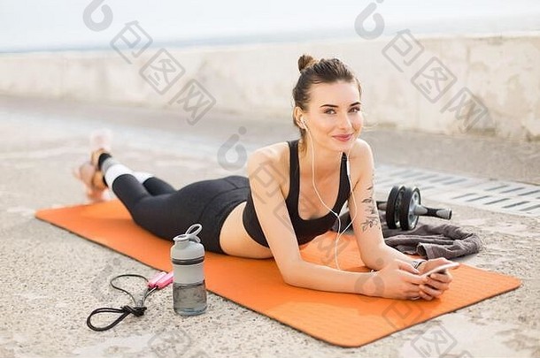 穿着<strong>运动</strong>上衣和带耳机的紧身裤的年轻美女躺在橙色瑜伽垫上，旁边放着<strong>运动</strong>瓶和跳绳，一边愉快地看着里面