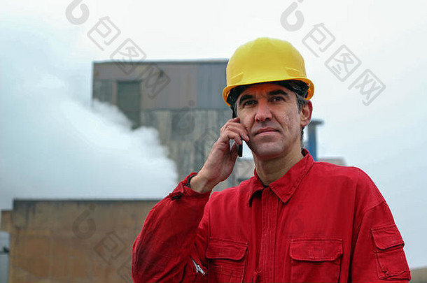 吸烟的工厂烟囱前，一名身穿红色工作服、戴黄色头盔、手持手机的<strong>工人</strong>的肖像。