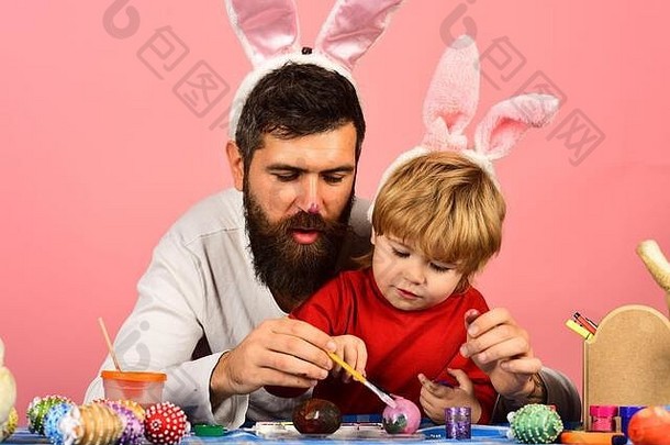 父亲和儿子正在为假期做准备。留着胡子的男人和小男孩在粉色背景上画复活节彩蛋。画鼻子的爸爸戴着兔子耳朵。复活节庆典和欢乐概念。