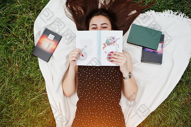 一位身穿黑色圆点连衣裙的美丽女子躺在草地上的毯子上，用一本书遮住脸的肖像。
