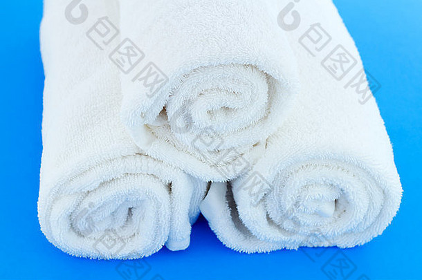 三块大的白色棉质浴布单独卷起，在蓝色背景上叠在一起。几条白色的棉质浴巾卷在蓝色的地板上。
