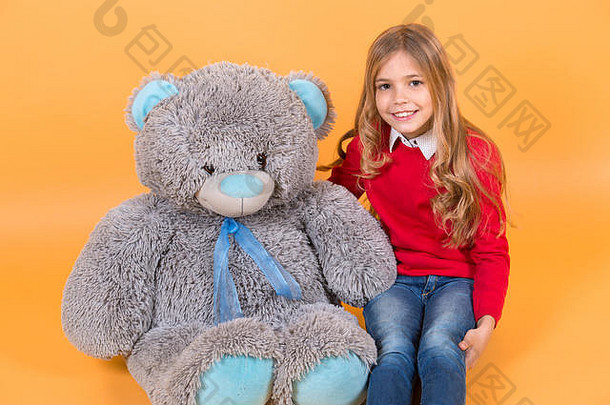 孩子带着灰色的软玩具微笑。女孩和橙色背景的大泰迪熊坐在一起。带动物娃娃的孩子，礼物和礼物。节日、生日、<strong>周年庆</strong>典。快乐童年的概念。