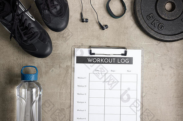 特写镜头：地板上的黑色运动鞋、耳机、健身跟踪器、一瓶水、负重板和剪贴板上的训练日志。